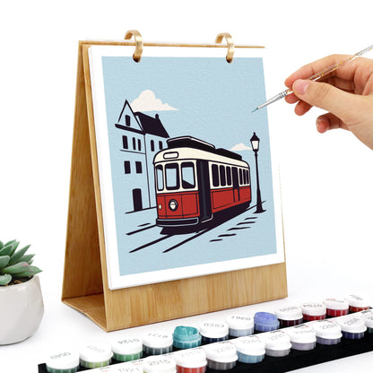 A wooden stand holding a colorful city scene painting with a red tram, white clouds, and buildings. A hand holds a fine paint brush near the artwork, with small round paint pots arranged along the bottom and a small potted succulent on the left.