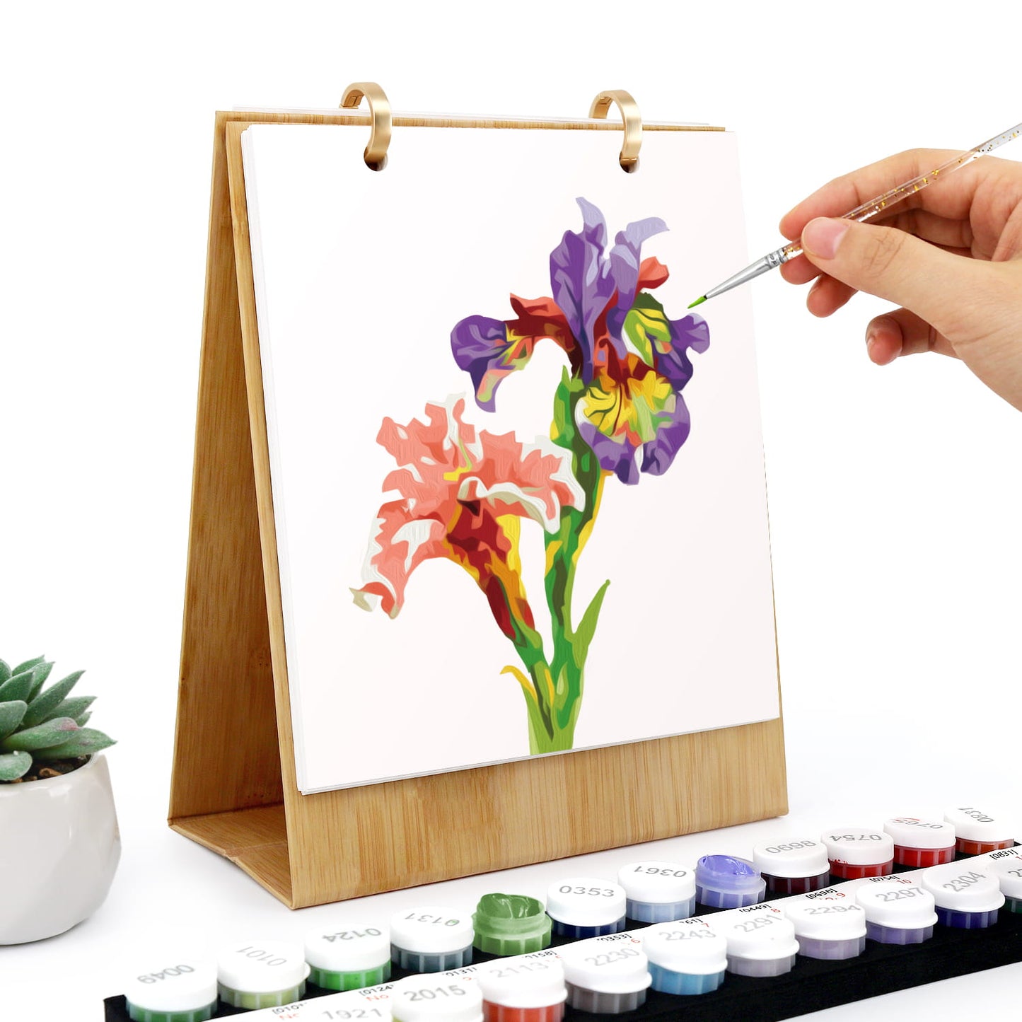 A hand is painting a colorful floral design on a wooden-framed canvas stand. The stand has a bamboo wood frame and gold metal rings at the top.