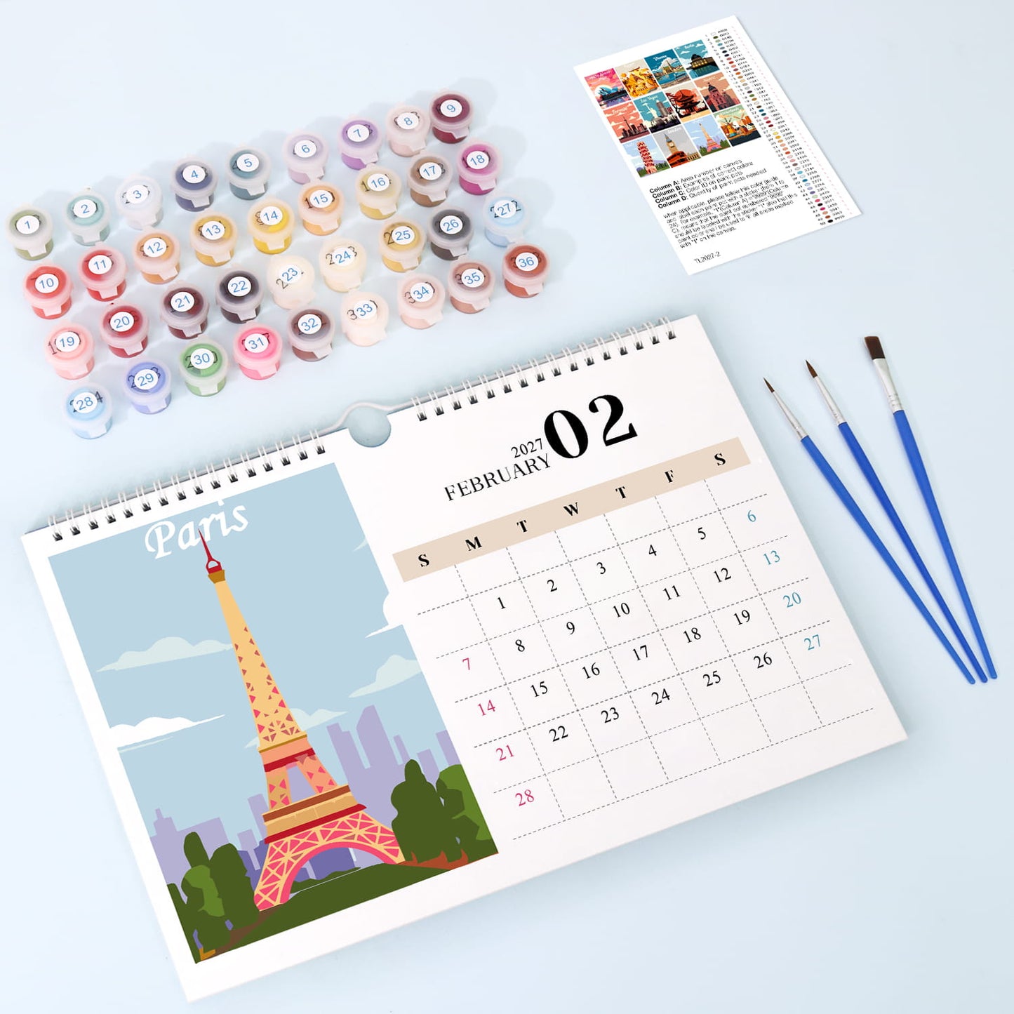 A flat-lay view of a Paris-themed desk calendar showing February 02, with a colorful Eiffel Tower illustration. Above the calendar are multiple small round paint pots in assorted colors, a small instruction card, and three blue-handled paintbrushes on a pale blue background.