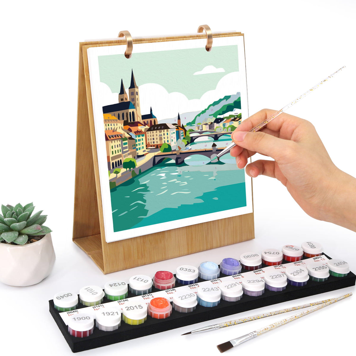 
A hand is painting on a wooden-framed canvas print of a colorful city river scene. In front is a tray of labeled paint pots and two paintbrushes, with a small potted succulent to the left, all on a white background.