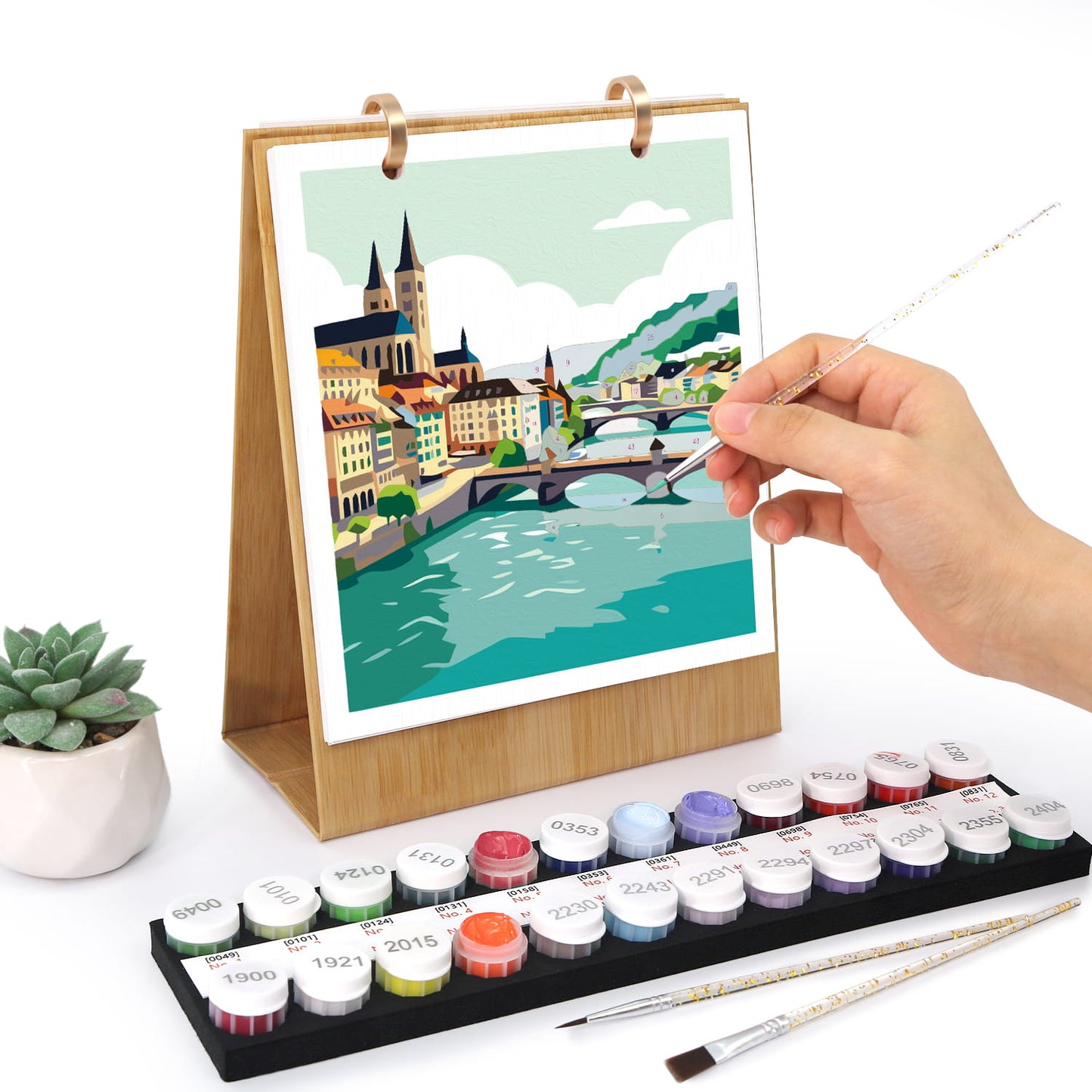 A hand is painting on a wooden-framed canvas print of a colorful city river scene. In front is a tray of labeled paint pots and two paintbrushes, with a small potted succulent to the left, all on a white background.