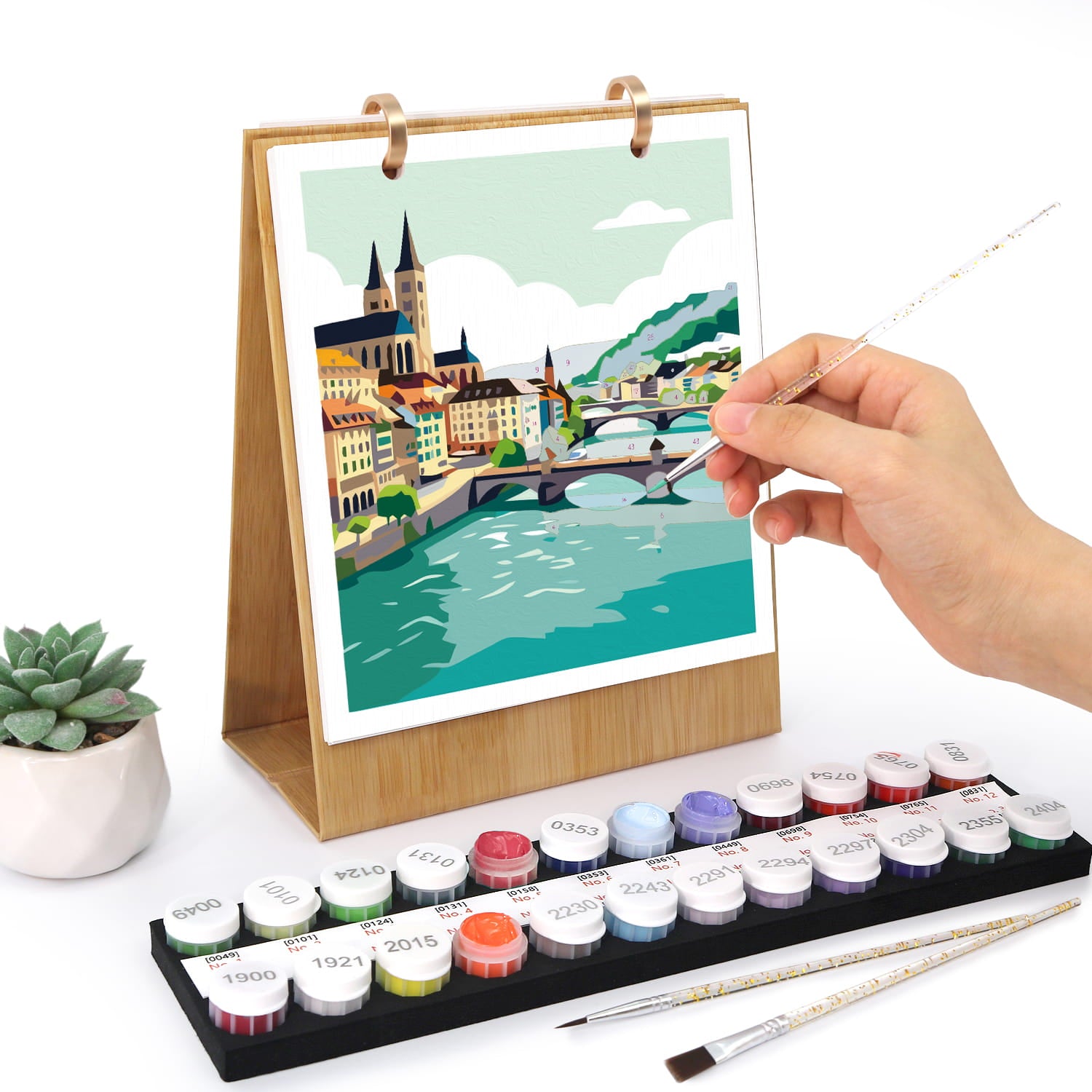 A hand is painting on a wooden-framed canvas print of a colorful city river scene. In front is a tray of labeled paint pots and two paintbrushes, with a small potted succulent to the left, all on a white background.