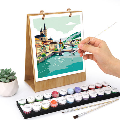 A hand is painting on a wooden-framed canvas print of a colorful city river scene. In front is a tray of labeled paint pots and two paintbrushes, with a small potted succulent to the left, all on a white background.
