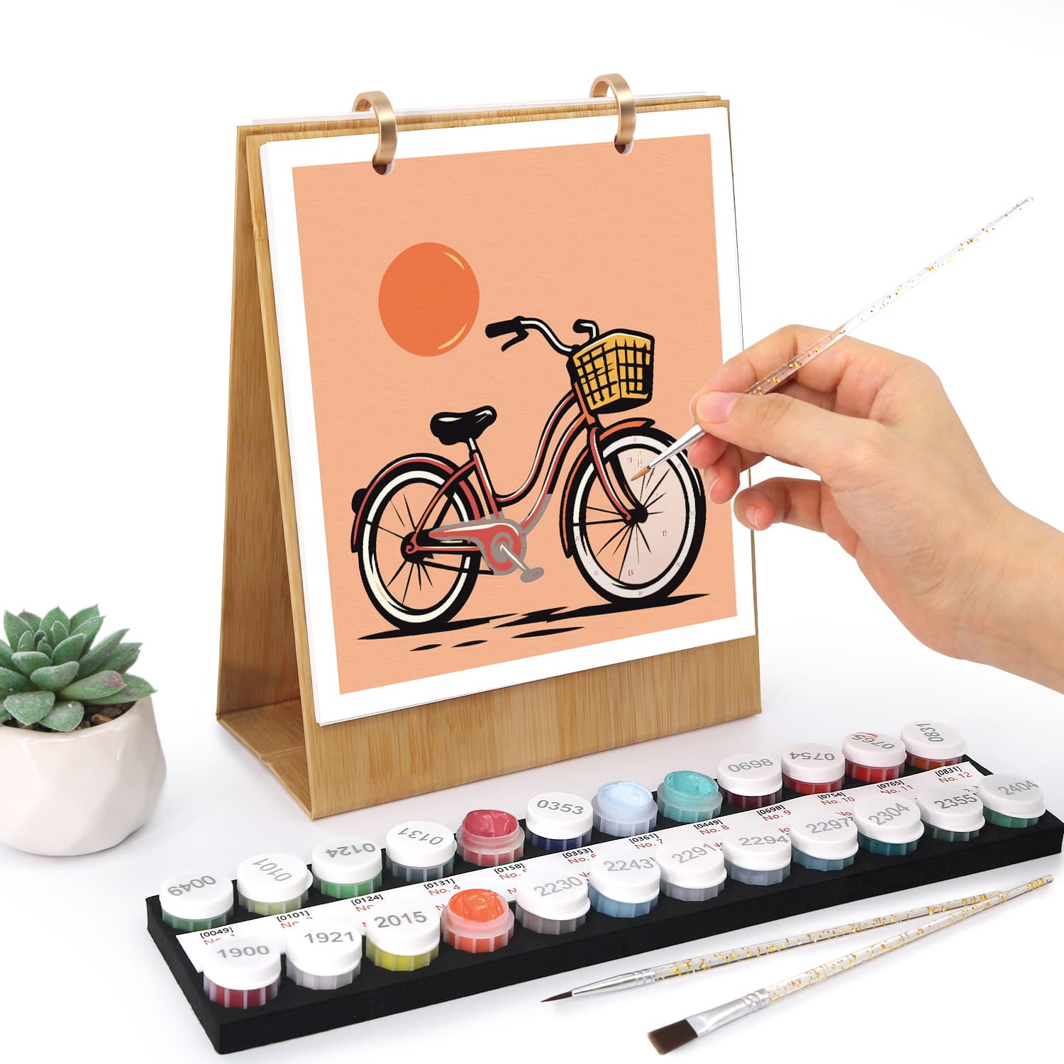 A hand is painting a bicycle illustration on a wooden-framed canvas stand, with a set of colorful paint tubes, two paintbrushes, and a small potted succulent on a white background.