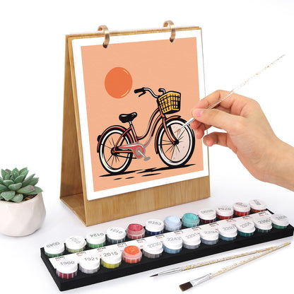 A hand is painting a bicycle illustration on a wooden-framed canvas stand, with a set of colorful paint tubes, two paintbrushes, and a small potted succulent on a white background.