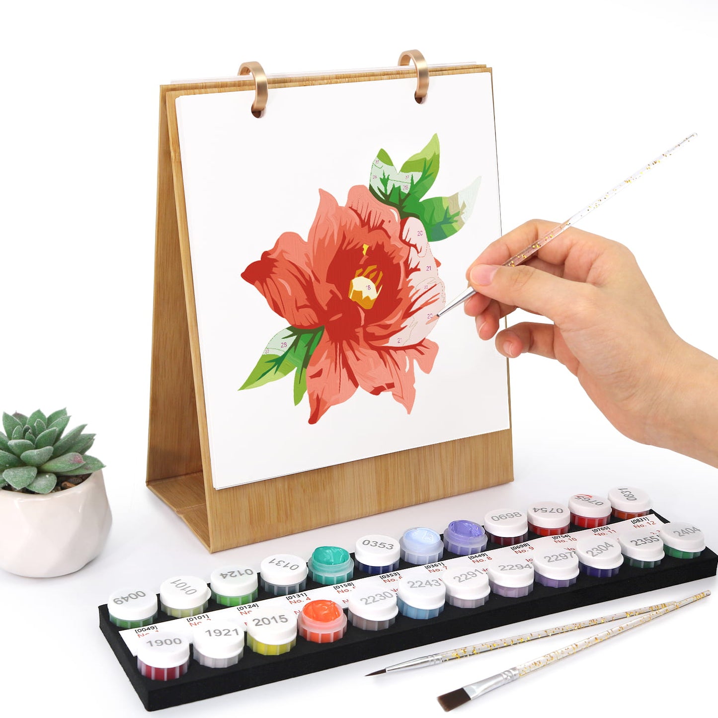 A hand holds a paintbrush over a small wooden easel displaying a painted red flower with green leaves. In front is a set of labeled paint pots and two paintbrushes on a white background, with a small potted succulent to the left.