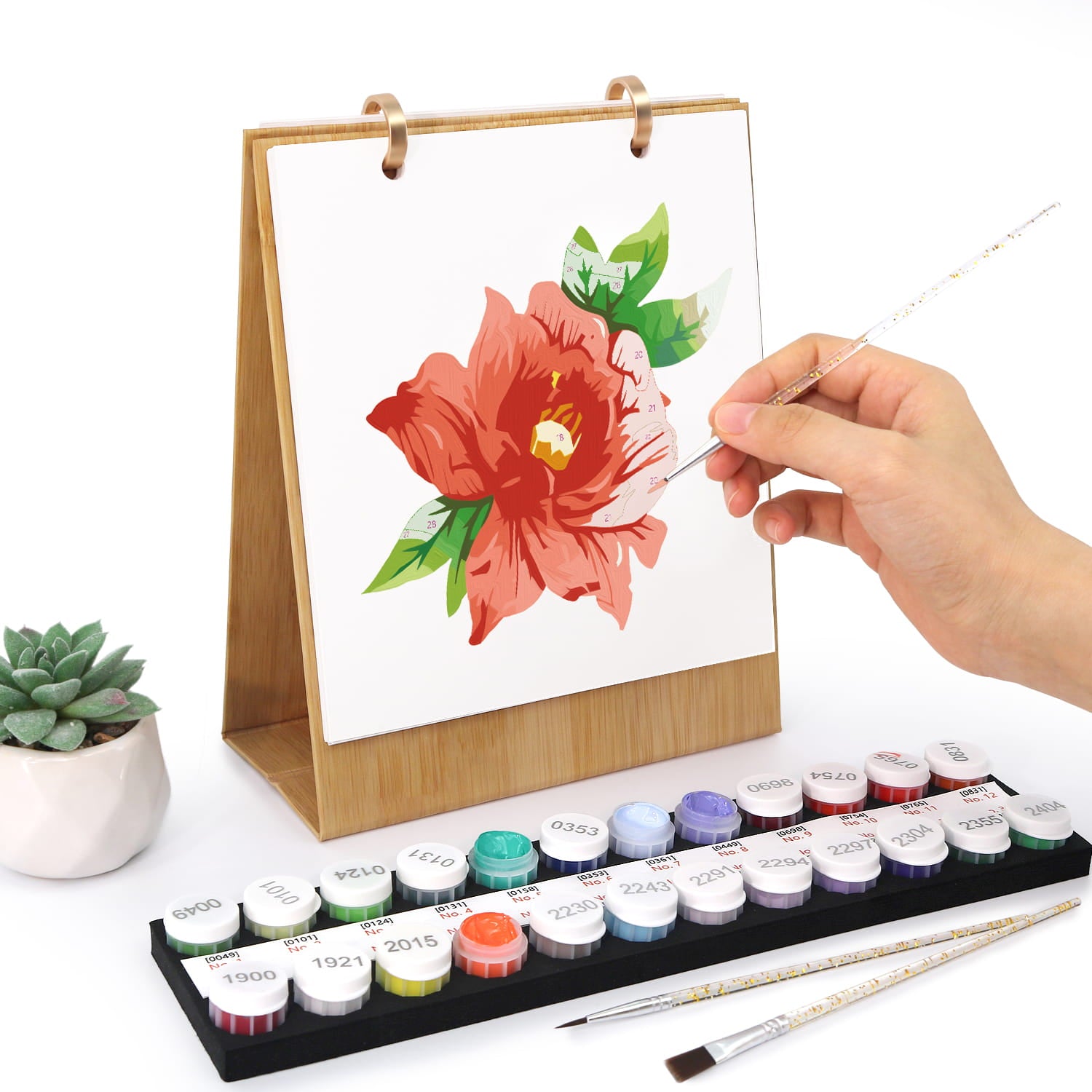 A hand holds a paintbrush over a small wooden easel displaying a painted red flower with green leaves. In front is a set of labeled paint pots and two paintbrushes on a white background, with a small potted succulent to the left.