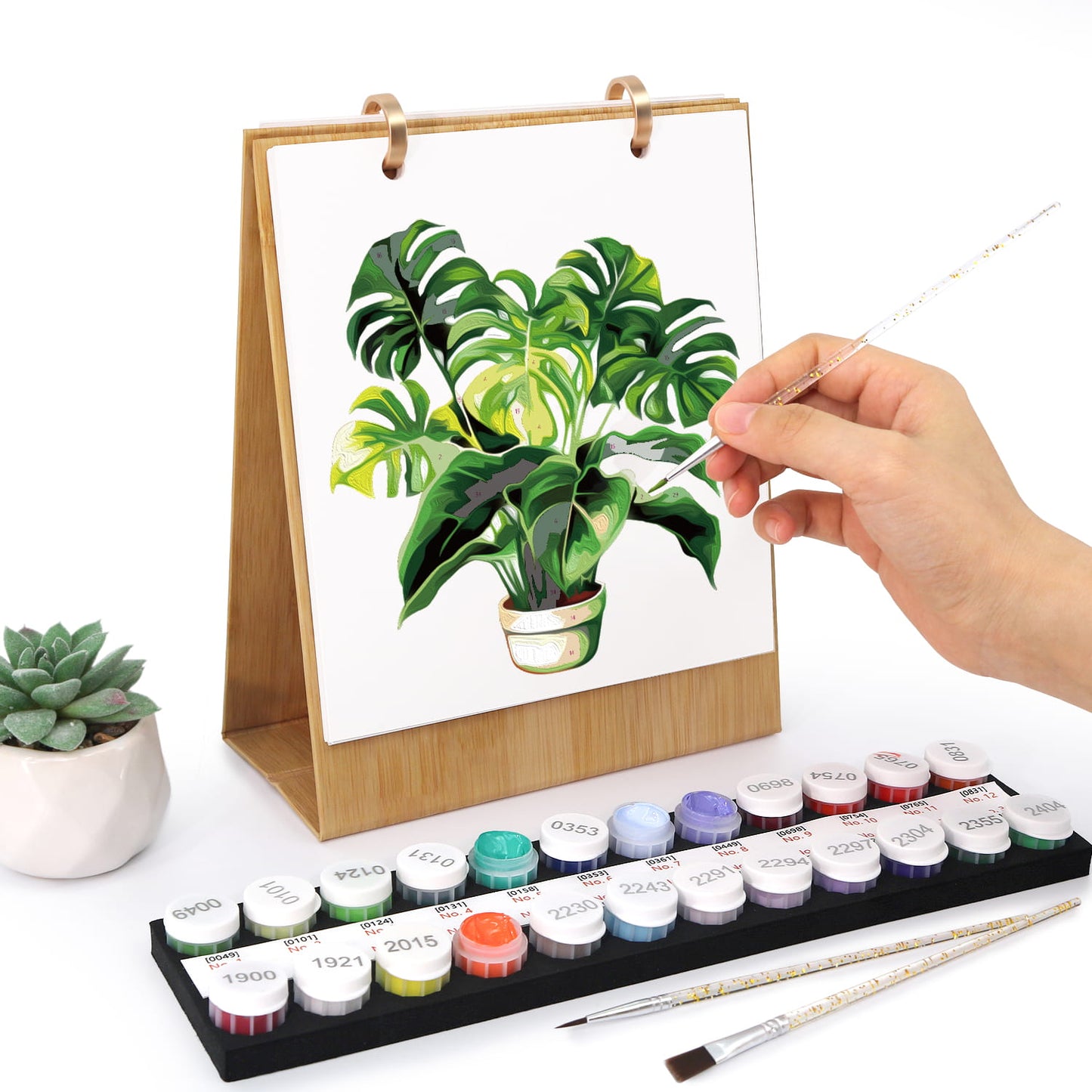A hand holds a paintbrush over a canvas showing a painted monstera plant, with a wooden easel stand behind it and a set of small paint tubes and brushes in front on a white background.