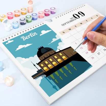 A spiral-bound desk calendar showing September 09 with a colorful illustration of a domed building and the word Berlin, photographed on a light blue surface. A hand holds a paintbrush over the calendar, and small paint pots are arranged around it.