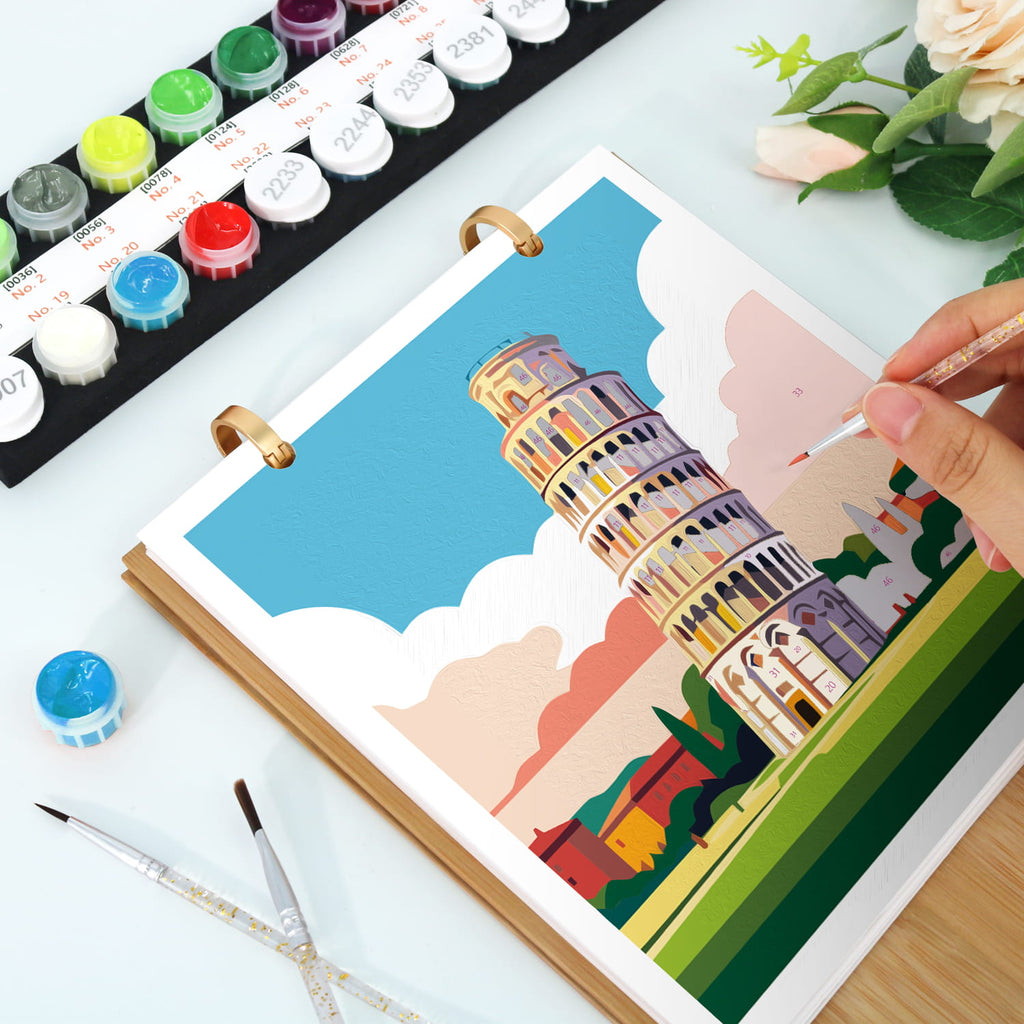 A hand is painting the Leaning Tower of Pisa in a ring-bound watercolor or paint-by-numbers style sketchbook. Paint tubes, paintbrushes, and flowers are arranged on a white tabletop.