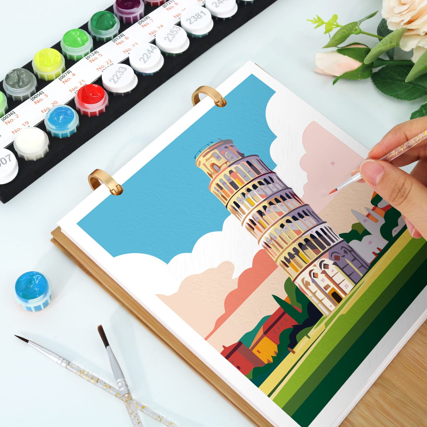 A hand is painting the Leaning Tower of Pisa in a ring-bound watercolor or paint-by-numbers style sketchbook. Paint tubes, paintbrushes, and flowers are arranged on a white tabletop.