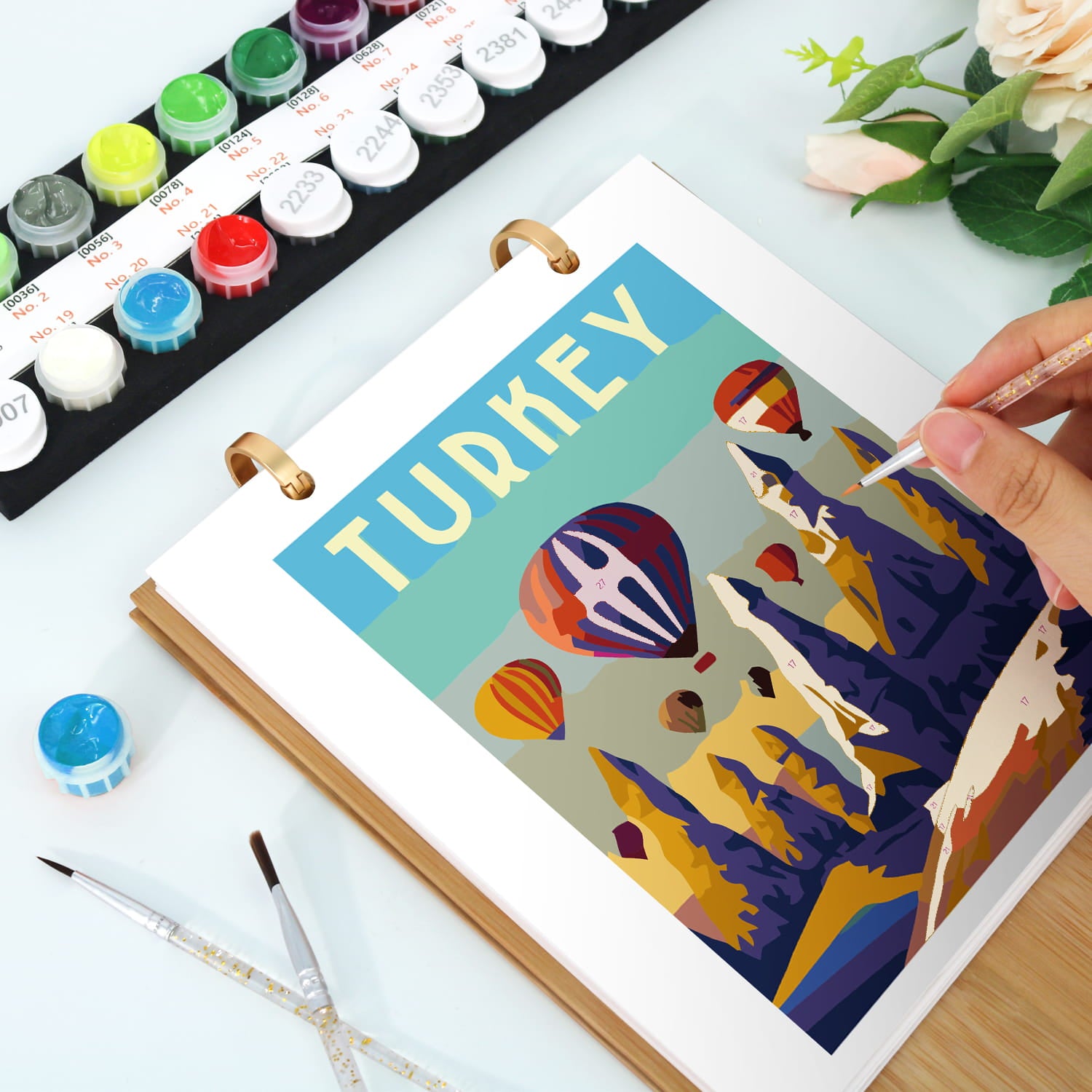A hand is painting on a ring-bound art print featuring a colorful stylized illustration of hot air balloons and mountains with the word 'TURKEY' in large letters. The scene is styled on a white surface with watercolor paints, paint tubes, and flowers in the background.