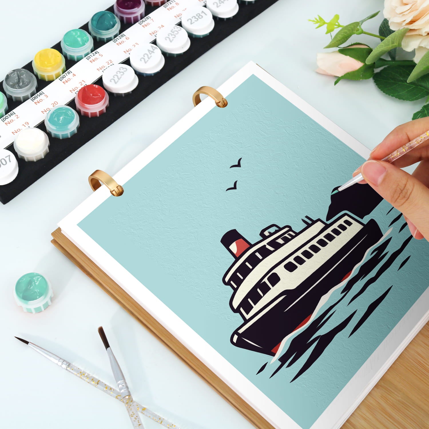 A hand is drawing a black and white ferry boat on a light blue canvas inside a spiral-bound art journal. The scene is styled as a desk with paint tubes, paintbrushes, and flowers.
