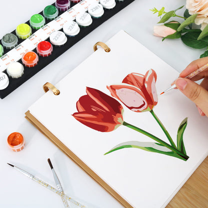 A hand is painting two red tulips on a white sketchbook with gold rings, with watercolor paint pots and paintbrushes visible on a white desk.