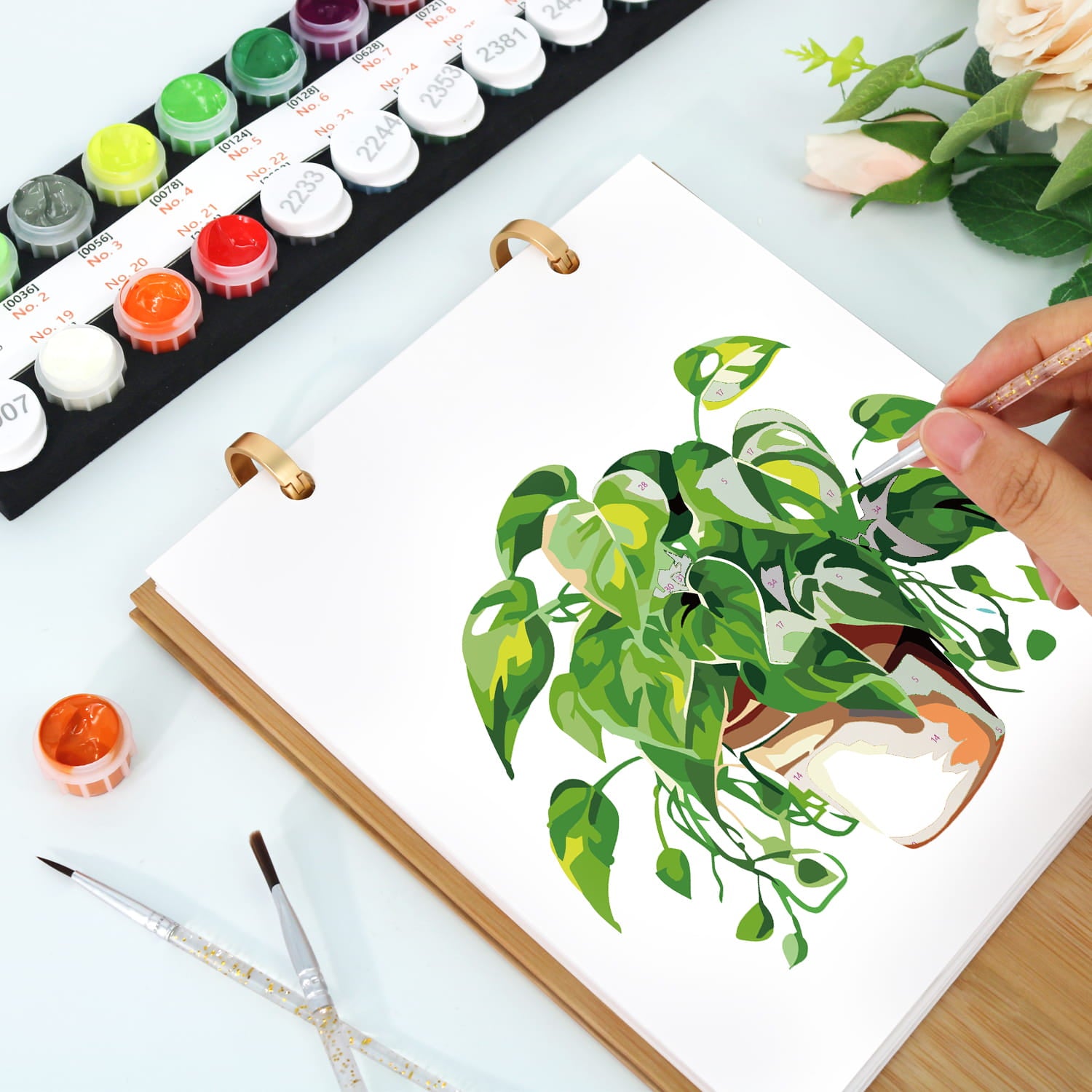 A hand is painting a green potted plant in a white ring-bound sketchbook, with watercolor paint pots, paintbrushes, and flowers arranged on a light tabletop.