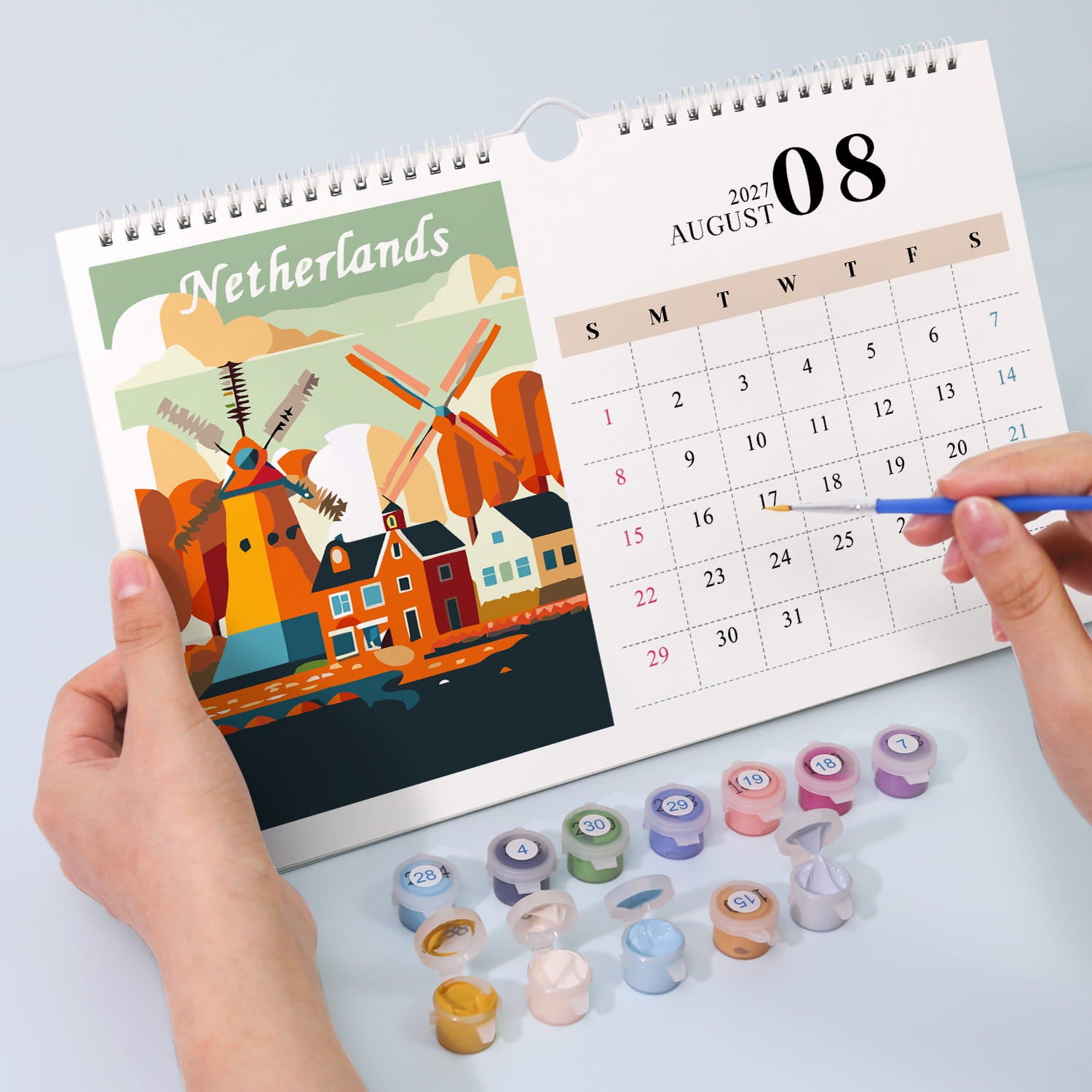 A spiral-bound Netherlands desk calendar is held open by two hands on a light gray surface. The left page shows a colorful illustration of Dutch windmills and houses with the word 'Netherlands' at the top.