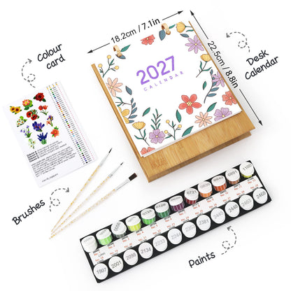 A flat-lay product presentation of a 2027 desk calendar kit, including the wooden-bound calendar with a floral cover, a color card, three paintbrushes, and a set of 24 watercolor paints in a black tray. The image features black text labels with arrows pointing to each component on a clean white background.