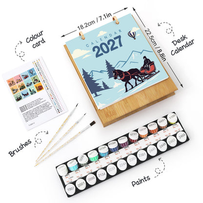A 2027 desk calendar set shown on a white background with dimension arrows and labels. The set includes a wooden-framed calendar with a winter horse-drawn carriage illustration, a colour card sheet, two paintbrushes, and a tray of small round paint pots.