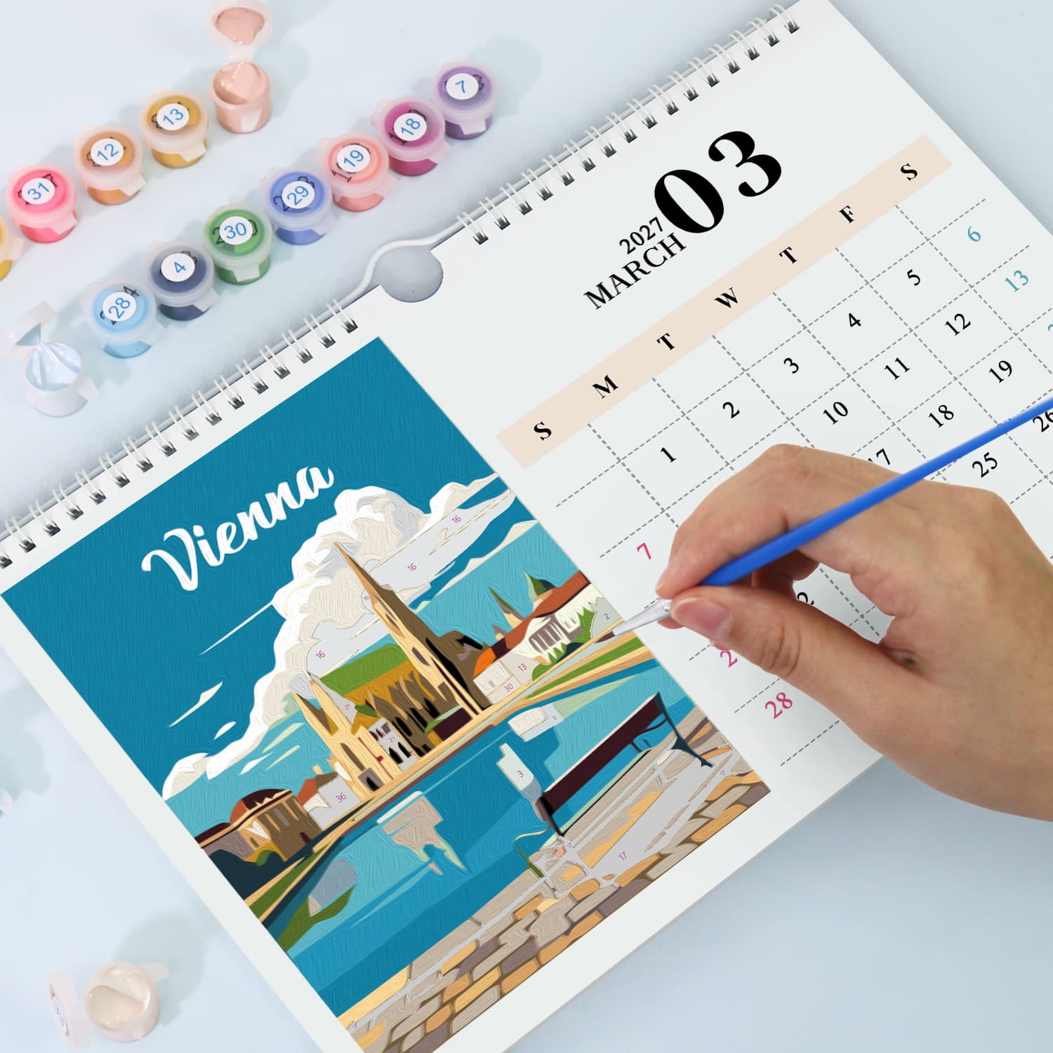 A spiral-bound desk calendar showing March 3, 2027, with a colorful illustrated cover featuring Vienna. A hand holds a blue pencil over the page, and small paint pots are arranged around it on a light surface.