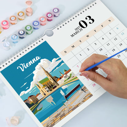 A spiral-bound desk calendar showing March 3, 2027, with a colorful illustrated cover featuring Vienna. A hand holds a blue pencil over the page, and small paint pots are arranged around it on a light surface.