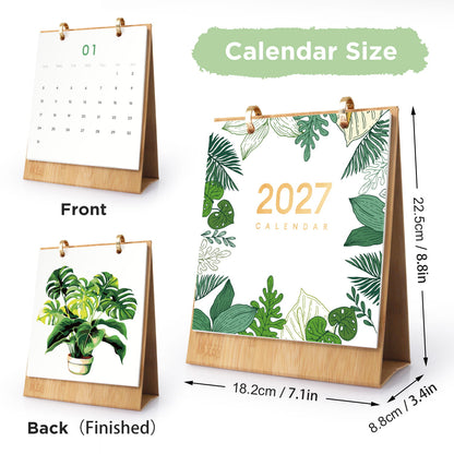 A desktop calendar shown in three views on a white background: front view, back view with a plant illustration, and a large front view with dimension arrows. The calendar has a light wood stand, gold-tone rings, and a white printed face with green tropical leaf illustrations and the text '2027 CALENDAR.
