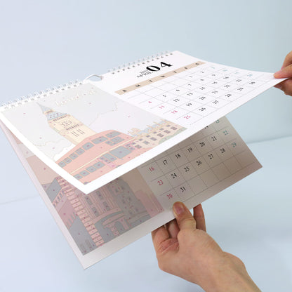 A spiral-bound desk calendar being held open by two hands, showing a printed cityscape page and a monthly calendar spread on a light blue background.