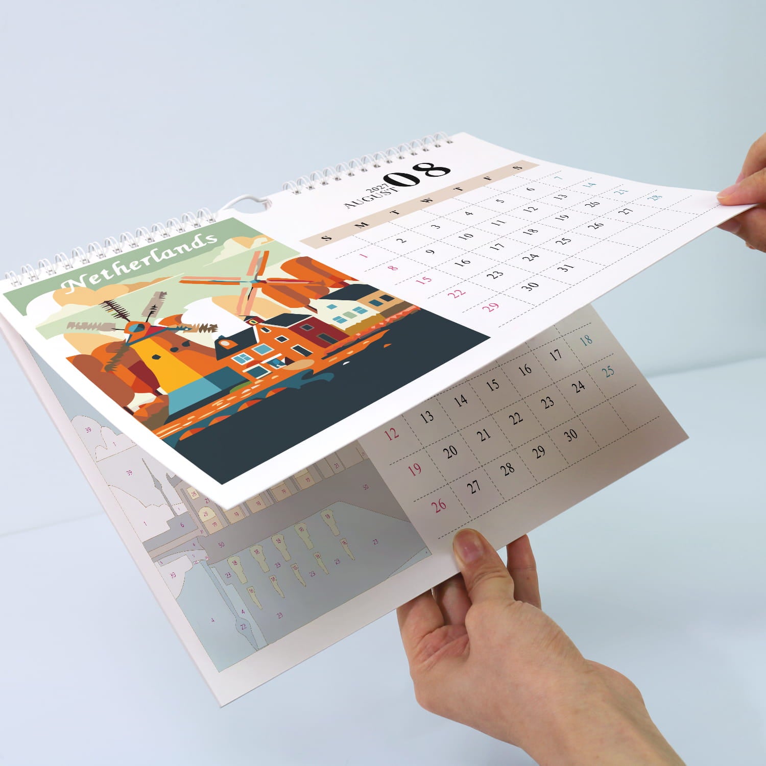 A spiral-bound desk calendar being held open by two hands, showing a Netherlands-themed page with colorful windmill artwork and a calendar grid for the 8th.