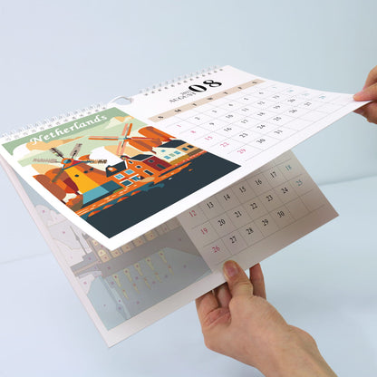 A spiral-bound desk calendar being held open by two hands, showing a Netherlands-themed page with colorful windmill artwork and a calendar grid for the 8th.