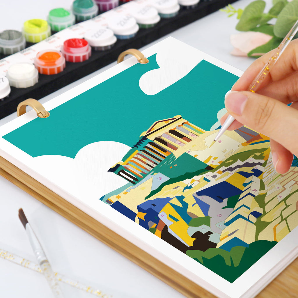 A hand holds a paintbrush over an open ring-bound art notebook showing a colorful geometric landscape illustration. The scene includes paint tubes, a palette, and a brush on a white surface.