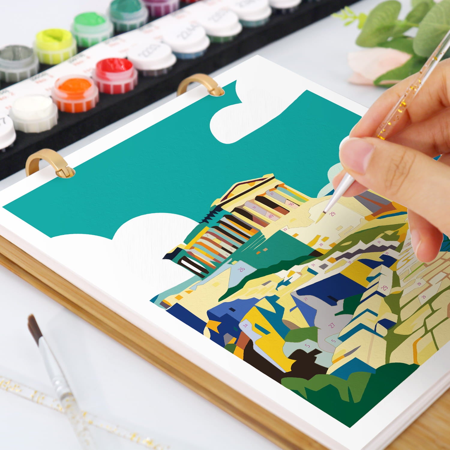 A hand holds a paintbrush over an open ring-bound art notebook showing a colorful geometric landscape illustration. The scene includes paint tubes, a palette, and a brush on a white surface.