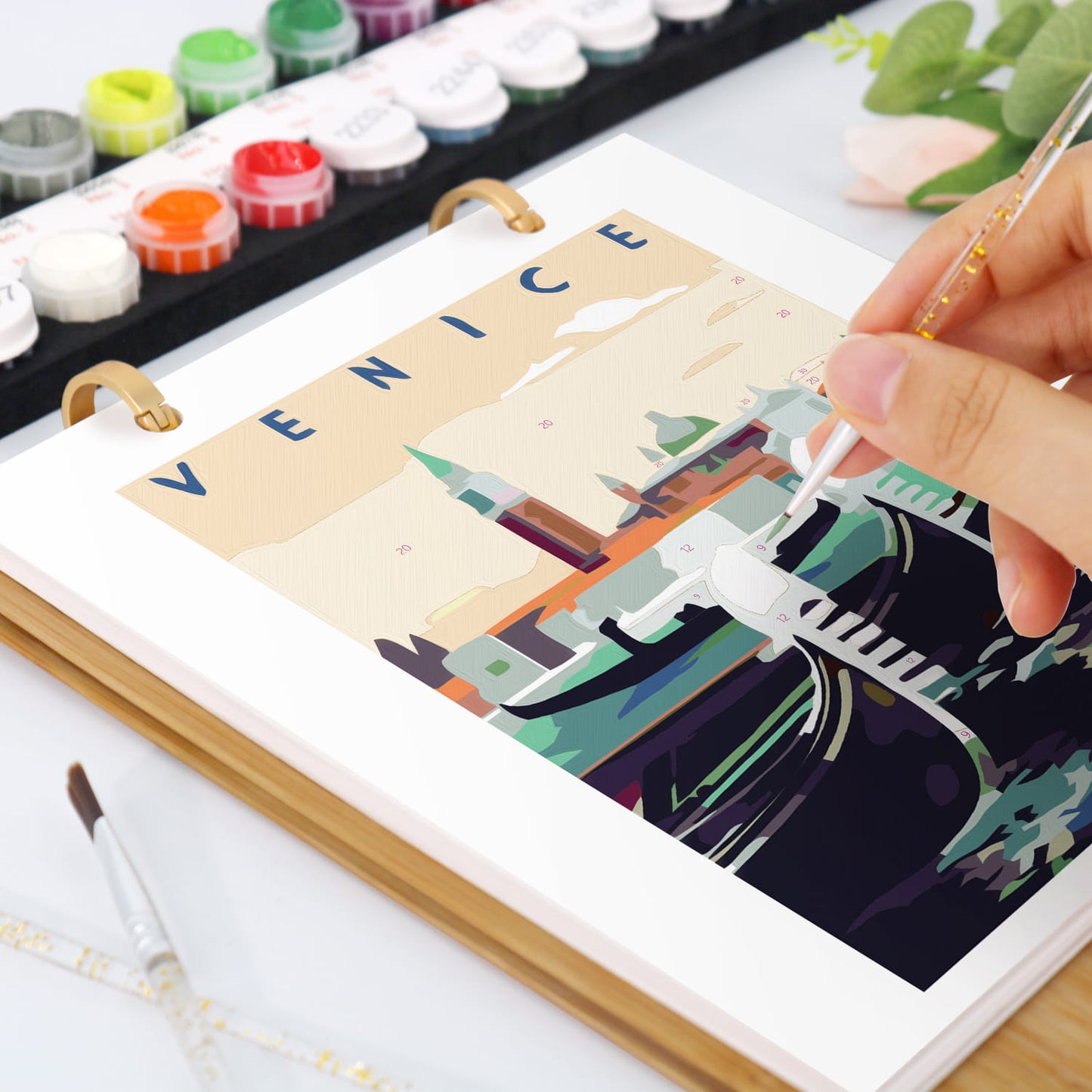A hand holds a paintbrush over an open art notebook showing a colorful Venice illustration. Paint tubes and a palette are visible in the background on a white surface.