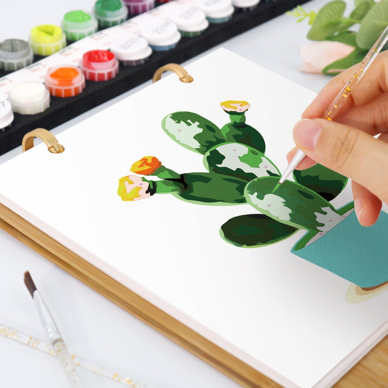 A close-up of a hand painting a green cactus with yellow and orange flowers in a white spiral-bound art notebook. Paintbrushes and paint tubes are visible on a white desk.