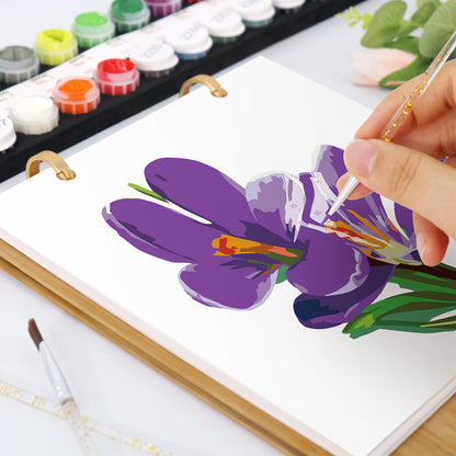 A hand painting a purple flower with green leaves on a white spiral-bound art notebook. Color paint pots are visible in the background.