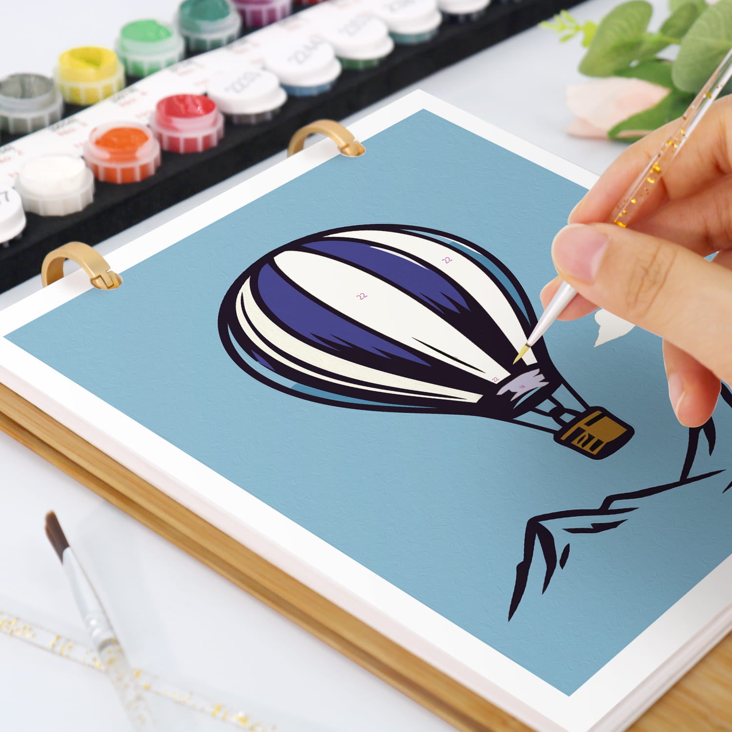 A hand is painting a hot air balloon illustration on a light blue sheet inside a wooden-bound art notebook. Paintbrushes, paint tubes, and a small plant are visible around it on a white surface.