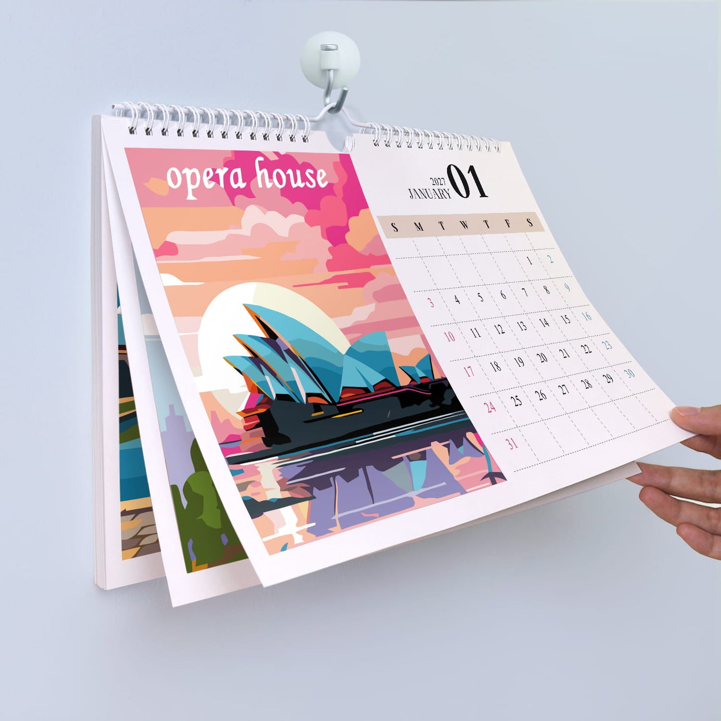 A spiral-bound desk calendar held open by a white clip against a light gray background. The front page features a colorful illustration of the Sydney Opera House with pink, orange, and blue tones, and the right page shows a January 2025 calendar grid.
