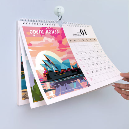 A spiral-bound desk calendar held open by a white clip against a light gray background. The front page features a colorful illustration of the Sydney Opera House with pink, orange, and blue tones, and the right page shows a January 2025 calendar grid.