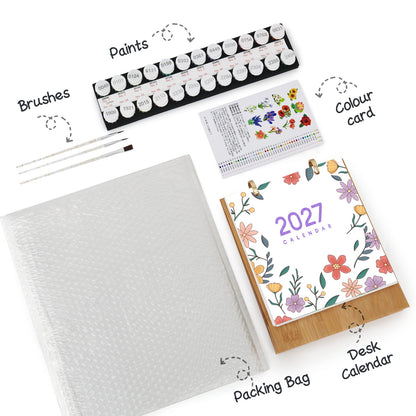 A flat-lay product shot of an art supplies and desk calendar set on a white background, with handwritten callouts pointing to each item: a tray of paint tubes, brushes, a colour card, a large white packing bag, and a 2027 floral desk calendar.