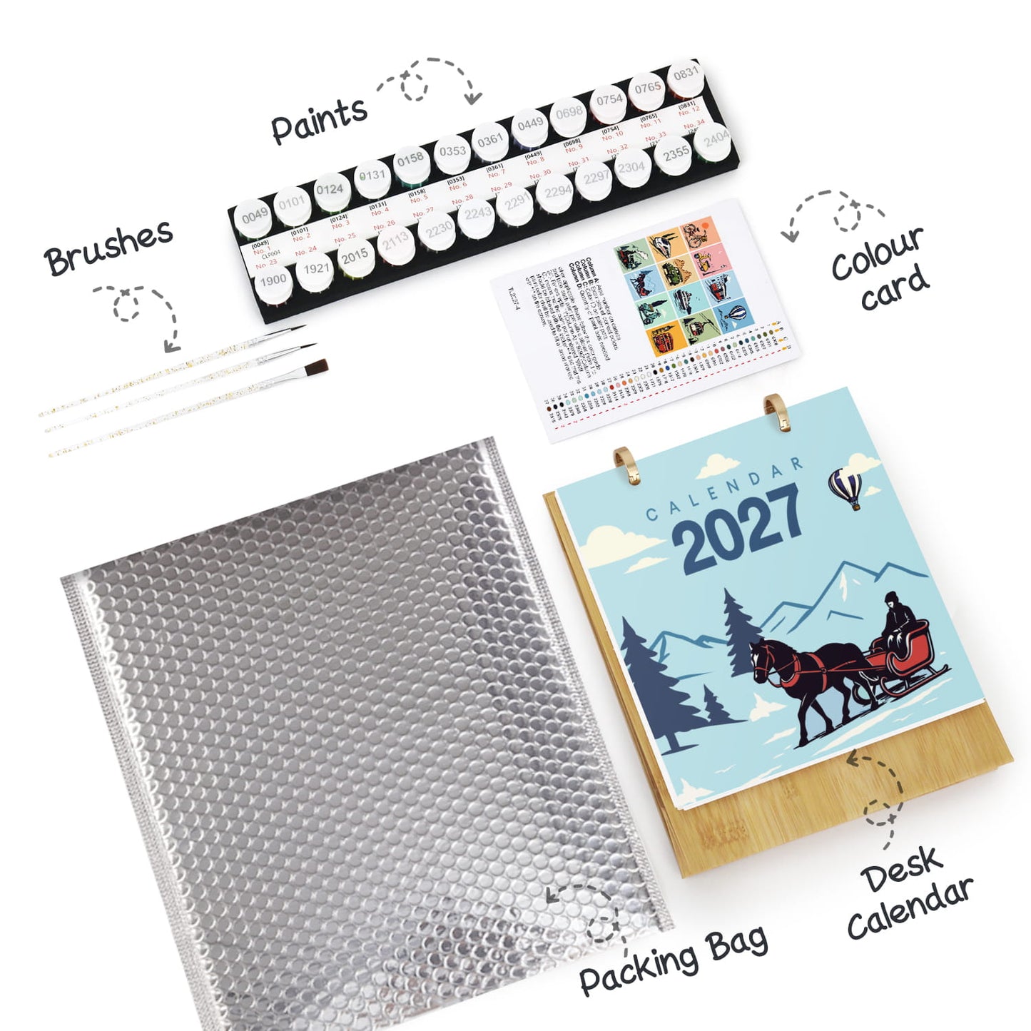 A flat-lay product shot of a children's painting set and desk calendar on a white background with handwritten callouts. The set includes a large silver bubble mailer packing bag, a black paint palette with round paint tubes, several white paintbrushes, a small color card, and a wooden desk calendar showing 2027 with a horse-drawn sleigh and mountain illustration.