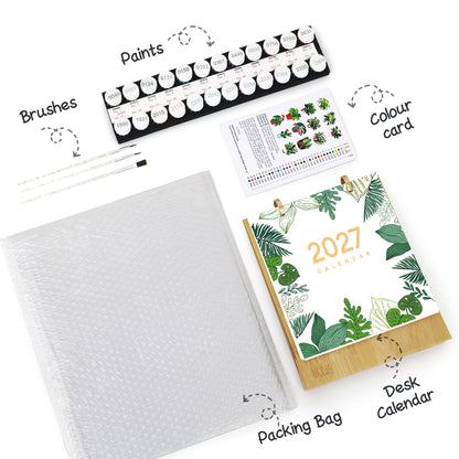 A flat-lay view of a 2027 desk calendar painting kit, showing the wooden desk calendar with a green tropical leaf pattern, a black paint palette with 12 paint tubes, a pack of brushes, a white textured packing bag, and a color card, all labeled with hand-drawn arrows and text on a white background.